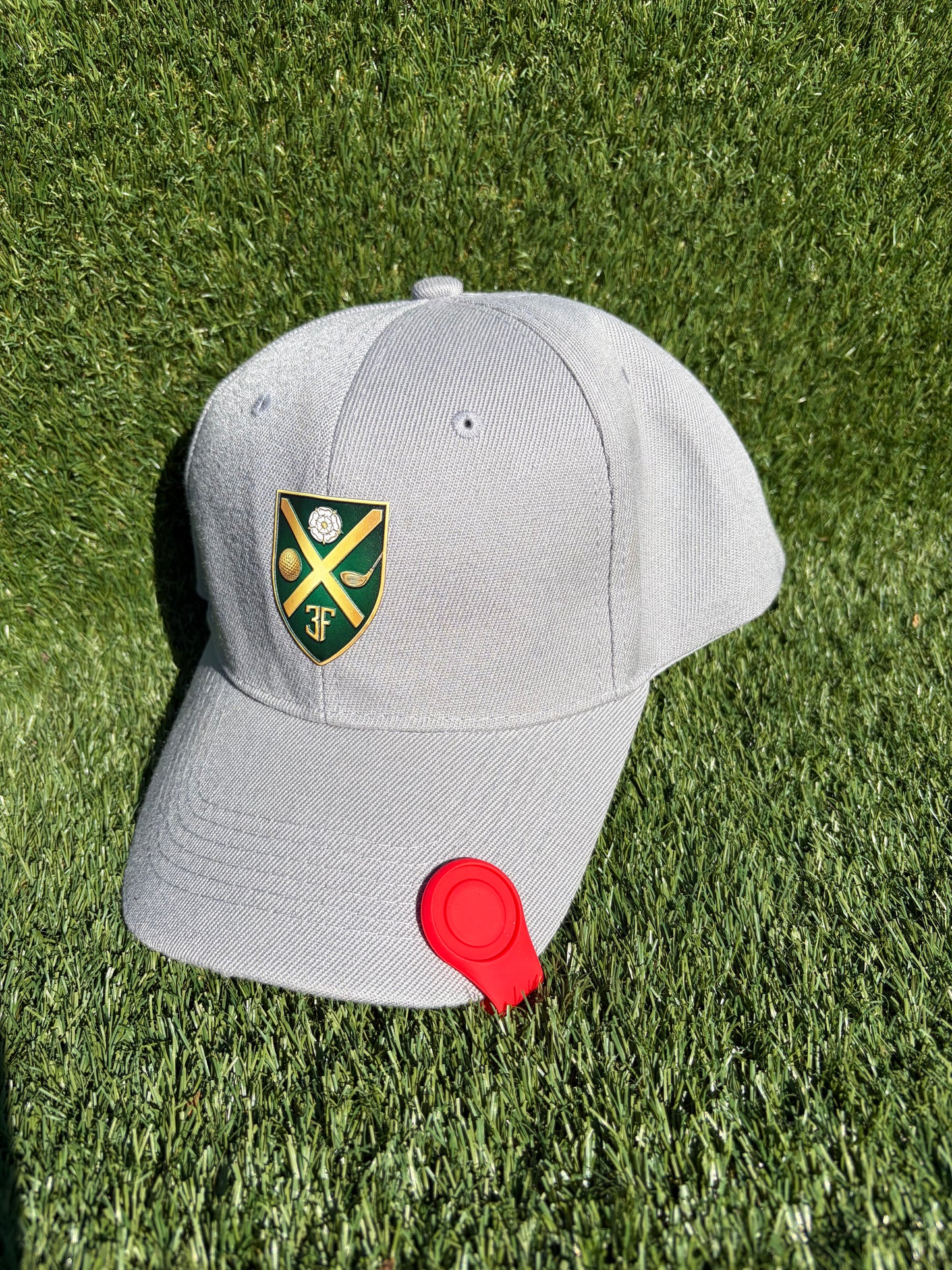 A light gray baseball cap with a green and yellow shield crest featuring crossed golf clubs and a golf ball, resting on green grass. A red silicone magnetic ball marker holder is attached to the brim.
