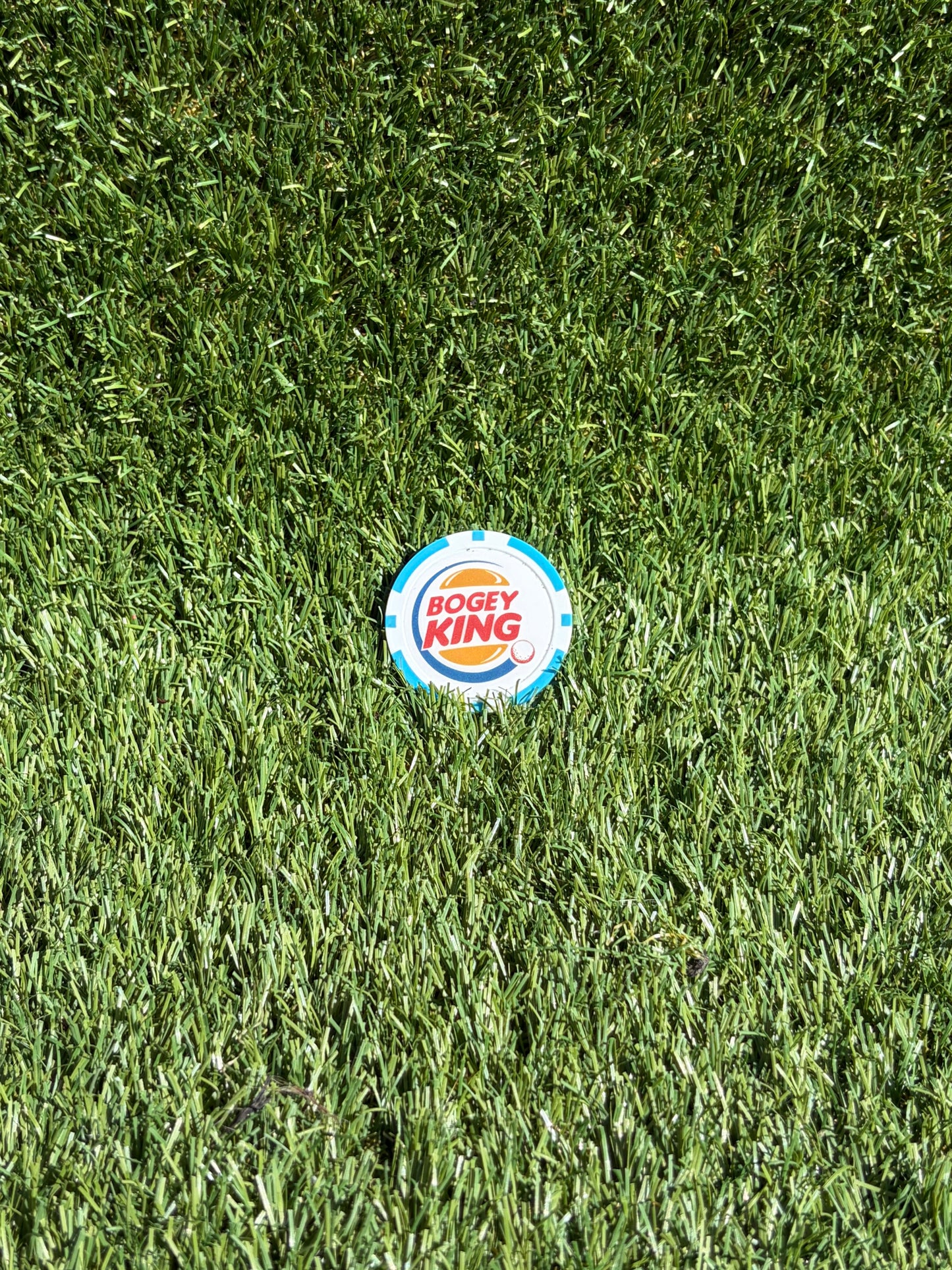 A circular Bogey King golf ball marker lies on a green grass surface.