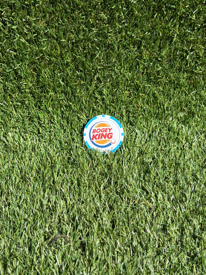 A circular Bogey King golf ball marker lies on a green grass surface.
