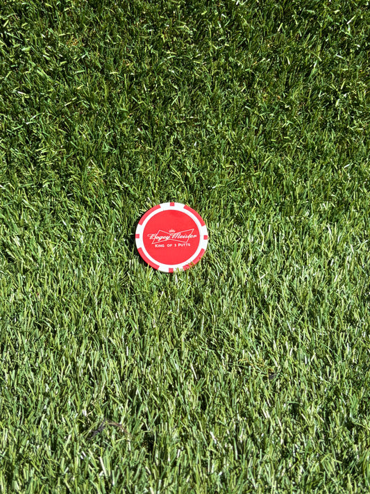 A single red and white golf ball poker chipball marker lies on a green grass surface, with the text 'bogey meister king of the 3 putts' visible on the chip.